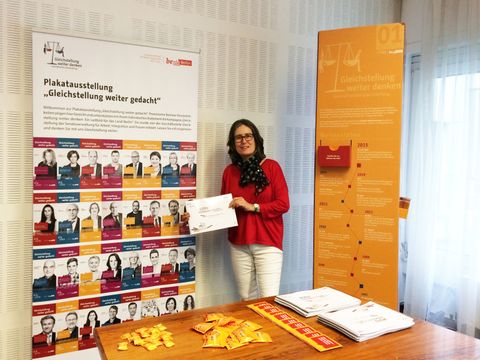 Bildvergrößerung: Frauenvertreterin Barbara Buse mit Memo-Malbuch neben Infostele und Plakat