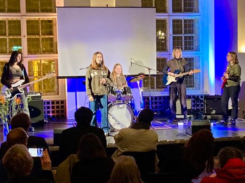 Bildvergrößerung: Eine Band steht auf einer Bühne. Viele Menschen sitzen auf Stühlen vor der Bühne.