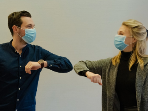 Eine Frau und ein Mann tragen eine medizinische Maske und begrüßen sich mit einem Ellbogencheck.