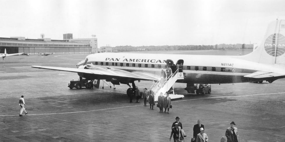 Eine schwarz-weiß Fotografie von einem Flugzeug auf einem Flughafen. Aus dem Flugzeug steigen Menschen aus.