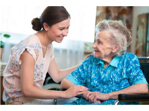 Junge Pflegerin sitzt lächelnd einer Seniorin gegenüber und streicht über deren Schulter