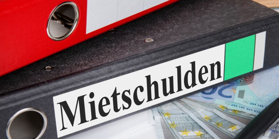 Ein Aktenordner mit der Aufschrift 'Mietschulden' liegt auf einem Schreibtisch. Auf ihm liegt ein weiterer Ordner, daneben ein Stift und mehrere Geldscheine.