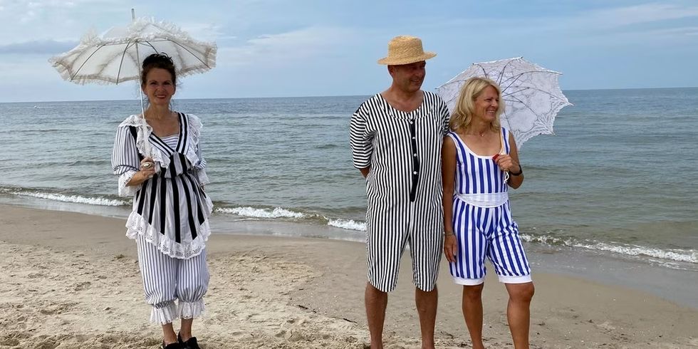 zwei Frauen mit Sonnenschirmen und ein Mann mit Hut in Badeanzügen aus der Gründerzeit am Strand