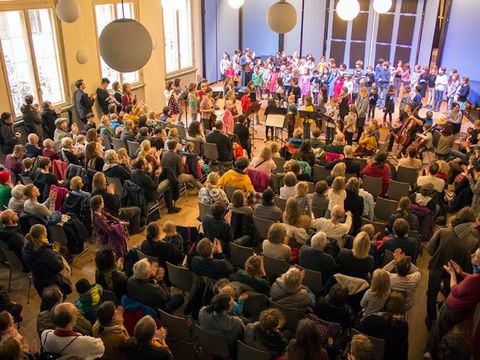 Voller Konzertsaal mit Bühne, darauf singende und musizierende Kinder