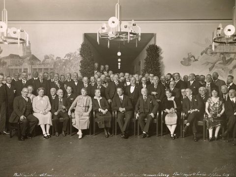 Bildvergrößerung: Schwarz-weiß-Fotografie von einer Gruppe Menschen, die in einem Saal posiert sitzen und stehen.