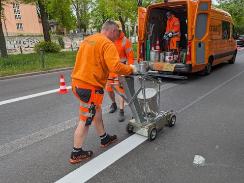 Bildvergrößerung: Ein Mann in oranger Arbeitskleidung zieht mit einem Gerät eine dicke weiße Linie über eine Straße.
