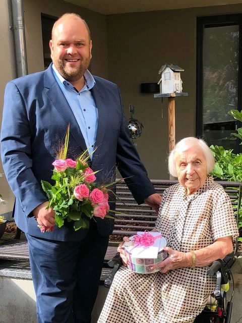 Bildvergrößerung: Ein Mann mit Blumenstrauß und eine ältere Frau im Rollstuhl mit Geschenk lächeln in die Kamera.