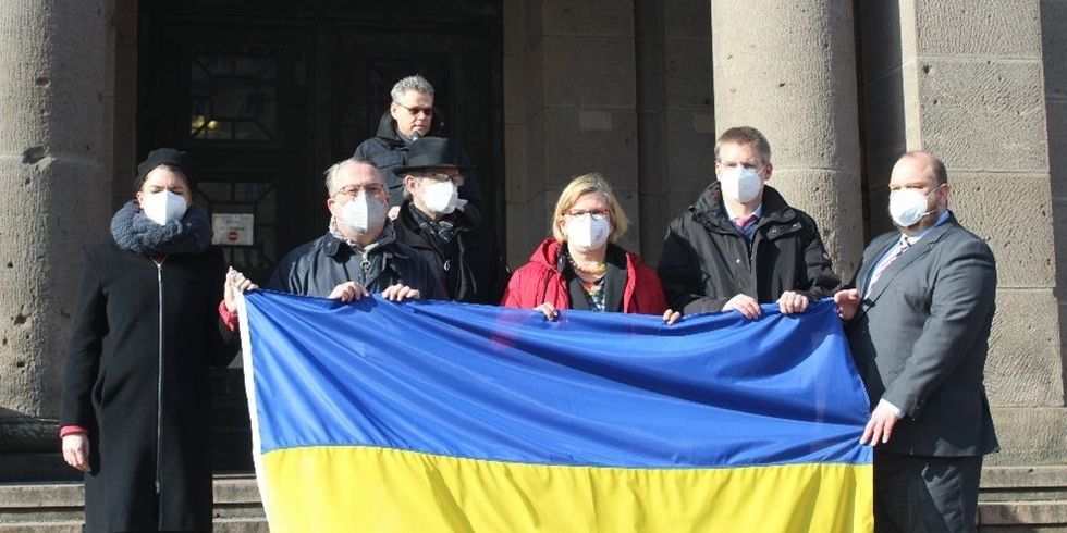 Sechs Personen stehen nebeneinander und halten zusammen die ukrainische Flagge. Ein Stück hinter ihnen steht eine weitere Person mit einem Mikrofon.