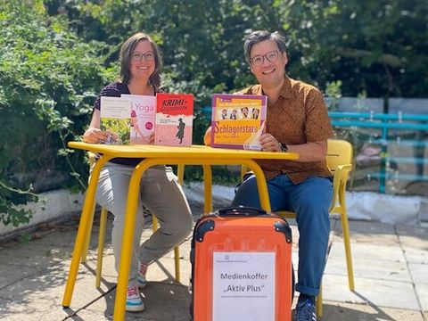 Bildvergrößerung: Frau und Mann sitzen an einem Tisch mit Büchern. Vor dem Tisch steht ein Koffer.