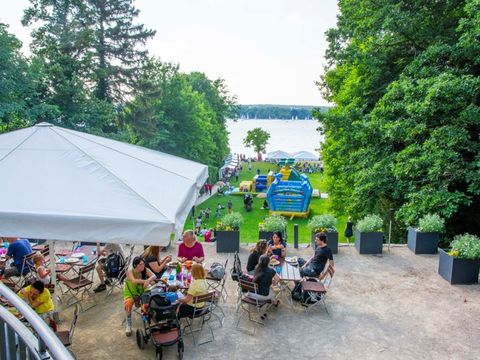 Auf einer Terasse an Tischen sitzen Menschen und unterhalten sich und essen. Hinter der Terasse liegt eine Grünfläche mit Hüpfburgen und noch hinter dieser Grünfläche erstreckt sich ein großer See.