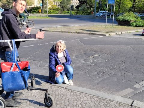 Bildvergrößerung: Auf dem Bild sind zwei Personen zu sehen. Die Person links ist ein Mann. Er hat braune Haare, er trägt einen schwarzen Pullover und eine dunkle Jeans und schaut ernst in die Kamera. Er nutzt eine Laufhilfe mit 2 großen Rädern hinten und 2 kleinen Rädern vorne. Eine blaurote Tasche hängt daran. Der Mann steht an einer Ampel, vor ihm liegt ein hoher Bordstein. Hinter dem Bordstein auf der Straße hockt eine Frau mit grauen Haaren. Sie trägt eine dunkelblaue Jacke und eine helle Jeans. Sie hält ein rundes rotes Schild hoch auf dem ein Daumen runter ist. Auch sie schaut ernst in die Kamera