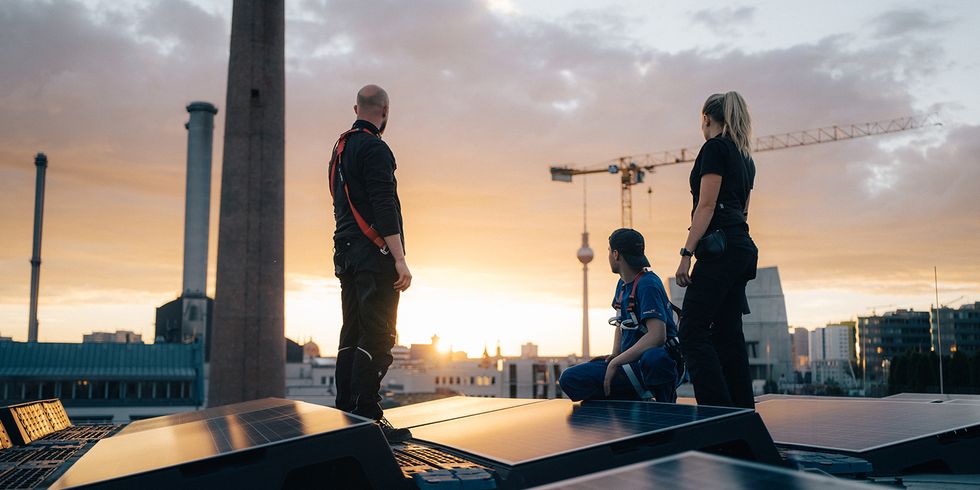 Drei Personen bei Sonnenaufgang auf dem Dach, im Vordergrund Solarpanel, im Hintergrund die Stadt Berlin.