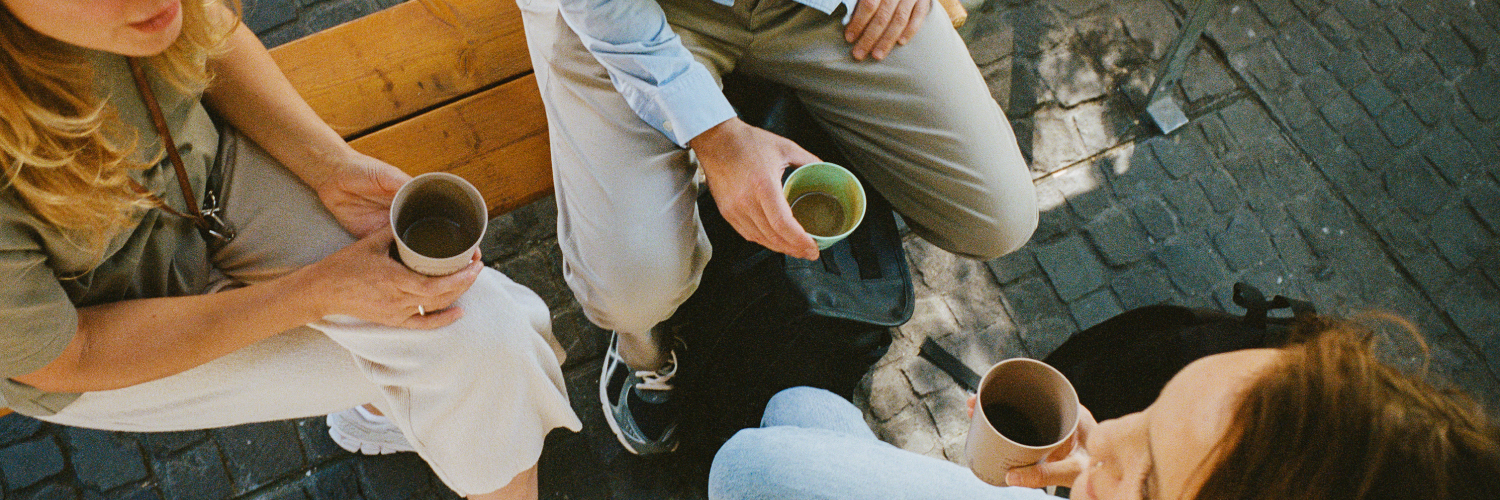 Drei Personen sitzen im Freien und halten Mehrweg-Kaffeebecher. Das Bild betont das Thema Mehrweg und nachhaltige Gewohnheiten im Alltag.