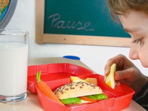 Ein Kind guckt lächelnd auf eine Brotdose und ein Glas Milch die auf einem Tisch stehen
