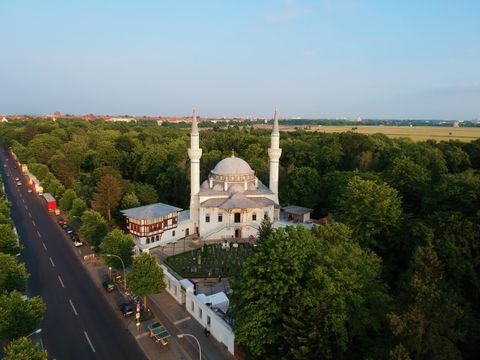Luftbildaufnahme der Moschee. Weißes Moschegebäude mit einer Kuppel in der Mitte. Rechts und links davon jeweils ein hoher, schlanker Turm. Die Moschee ist umgeben von vielen dunkelgrünen Bäumen 