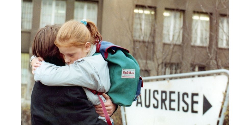 Kurt Naumann (Daniel Brenner, von hinten) umarmt Judith Richter (Johanna Brenner) zum Abschied, trägt sie im Arm; Richter (Johanna Brenner) trägt einen Rucksack; im Hintergrund Schild "Ausreise"