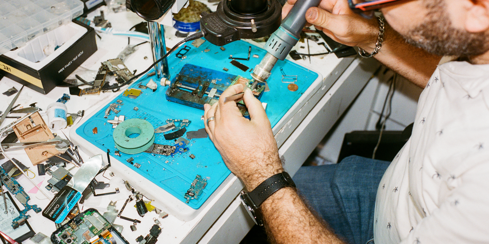 Eine Person repariert elektronische Geräte auf einer Werkbank, umgeben von Werkzeugen und Ersatzteilen. Sie nutzt einen Lötkolben, um kleine Bauteile zu bearbeiten. Das Bild zeigt die Bedeutung von Reparatur und Wiederverwendung im Sinne der Zero-Waste-Bewegung und fördert eine nachhaltige Nutzung von Elektrogeräten.