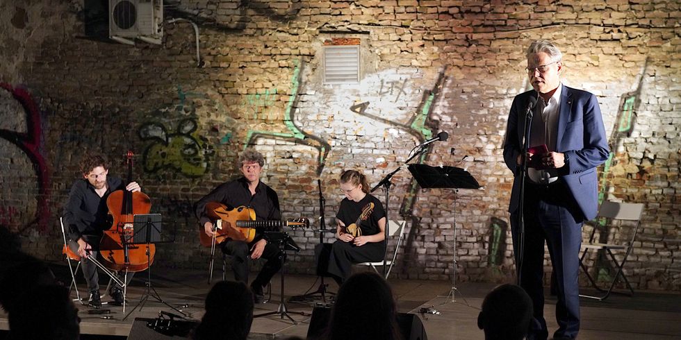 Ein Mann spricht draußen im Dunkeln auf einer beleuchteten Bühne vor Publikum in ein Mikrofon, im Hintergrund sitzen drei Musiker_innen an ihren Instrumenten.