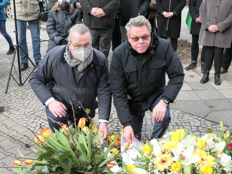 Bildvergrößerung: Zwei Männer hocken vor mehreren Blumensträußen. Im Hintergrund ist eine Menschenmenge.