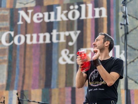 Im Vordergrund ein Mann mit einem schwarzen T-Shirt. Er hält einen Schellenkranz in der Hand mit dem er gerade Musik macht. Im Hintergrund der weiße Schriftzug "Neukölln Country & Folk Festival"
