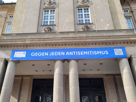 Bildvergrößerung: Vor einem großen Gebäude hängt über dem Eingang ein Banner mit der Aufschrift gegen jeden Antisemitismus.