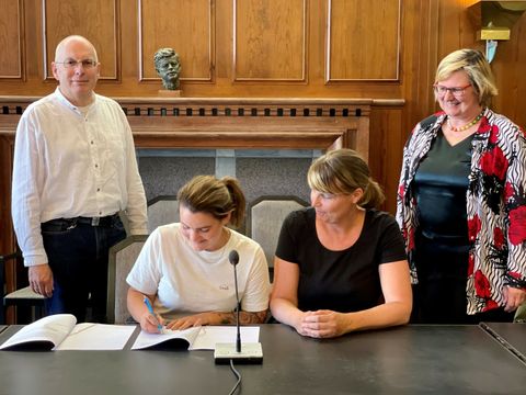 Bildvergrößerung: Zwei Frauen sitzen an einem Tisch. Eine der beiden Frauen unterschreibt auf einem Blatt Papier. Die andere schaut zu ihr rüber. Zwei weitere Menschen stehen neben den Frauen.