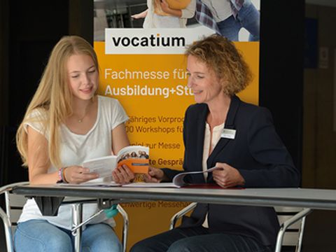 Zwei Frauen sitzen an einem Tisch vor dem Plakat der Fachmesse vocatium 