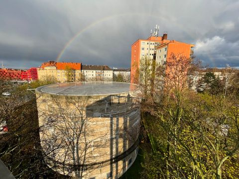 Bildvergrößerung: Ein großer Betonzylinder steht zwischen Bäumen. Im Hintergrund ist vor einem bewölkten Himmel ein Regenbogen zu erkennen.