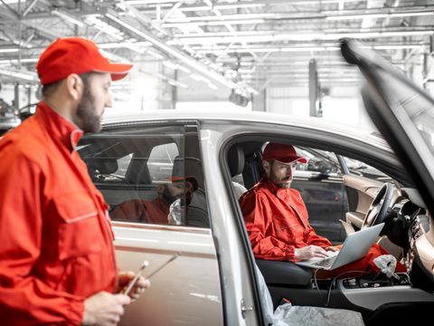 Zwei männliche Automechaniker in roter Uniform diagnostizieren Auto mit Computer beim Autoservice