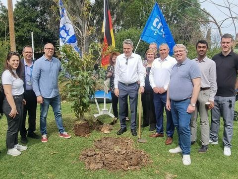 Bildvergrößerung: Ein Gruppe von Menschen steht auf einem Rasen, im Hintergrund die israelische und die deutsche Flagge. Ein Baum steht zwischen diesen Menschen einpflanzbereit neben einem frisch gegrabenen Loch im Rasen.