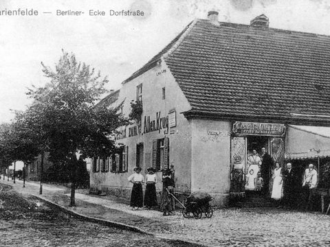 Bildvergrößerung: Eine schwarz-weiß Fotografie von einem Haus an einer Straße. Vor und neben dem Haus stehen Menschen.