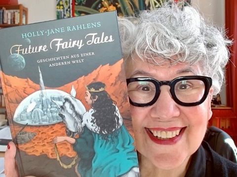 Bildvergrößerung: Eine Frau lächelt in die Kamera und hält neben ihr Gesicht ein Buch hoch. Das Buch ist Future Fairy Tales, Geschichten aus einer anderen Welt, von Holly-Jane Rahlens.