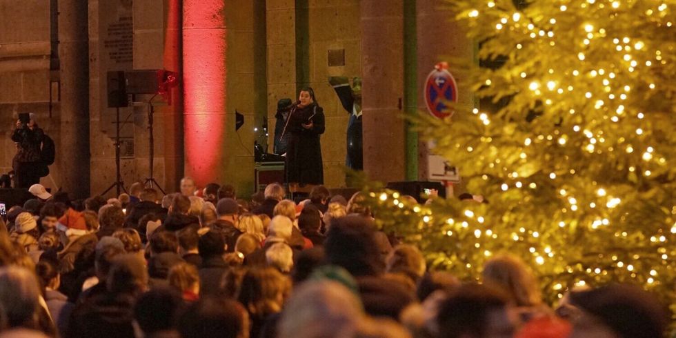 Eine Frau steht an einem Mikrofon vor einem großen Gebäude. Auf dem Platz davor stehen viele Menschen neben einem beleuchteten Weihnachtsbaum und schauen zu ihr hin.