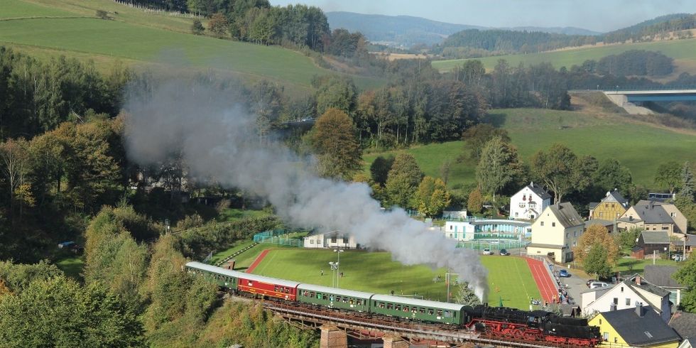 über eine Brücke fahrender Zug mit Dampflok zwischen Häusern und Wiesen