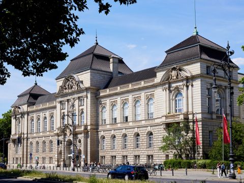 Das Bild zeigt das Gebäude der Universität der Künste in Berlin. Es handelt sich um ein imposantes, historisches Bauwerk mit einer reich verzierten Fassade aus hellem Stein und großen Fenstern. Das Dach ist dunkel und hat mehrere Türme. Vor dem Gebäude sind Straßen, Gehwege und einige parkende Autos zu sehen. Ein paar Bäume und rote Fahnen flankieren das Gebäude, und der Himmel ist klar und blau.