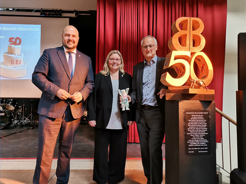 Bildvergrößerung: Bezirksbürgermeister Frank Bewig, Bildungsstadträtin Dr. Carola Brückner und Schulleiter Harald Schwarz auf der Bühne der Aula neben der anlässlich des Jubiläums enthüllten Skulptur „50 Jahre BBO“.