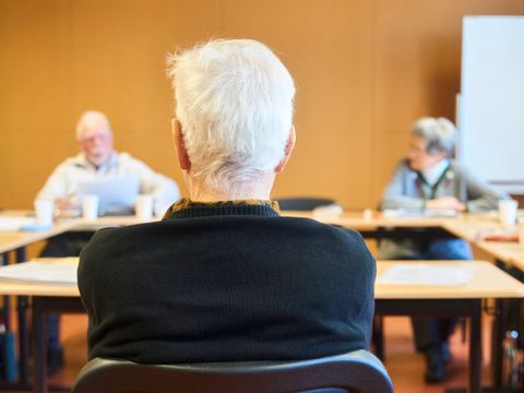 Schulterblick über Kursleitung im Unterricht. Kursleitung ist von hinten zu sehen, die Teilnehmenden unscharf im Hintergrund.