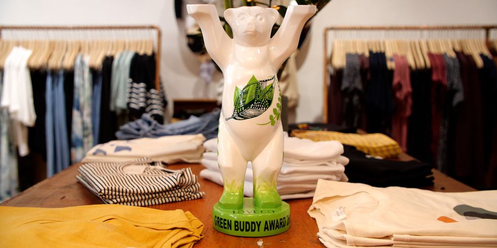 Eine Bärenfigur mit grünen Blättern und der Aufschrift "Green Buddy Award" steht auf einem Tisch mit zusammengelegten Kleidungsstücken. Im Hintergrund sind weitere Sachen an Stangen.