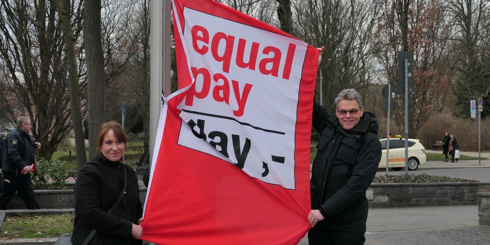 Eine Frau und ein Mann hissen eine Fahne mit der Aufschrift Equal Pay Day