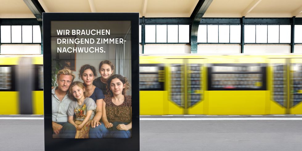 Das Bild zeigt eine Werbetafel auf einem Bahnsteig. Die Werbetafel zeigt eine Familie, die aus einem Mann, einer Frau und drei jungen Mädchen besteht. Auf der Tafel steht: “WIR BRAUCHEN DRINGEND ZIMMERNACHWUCHS.”