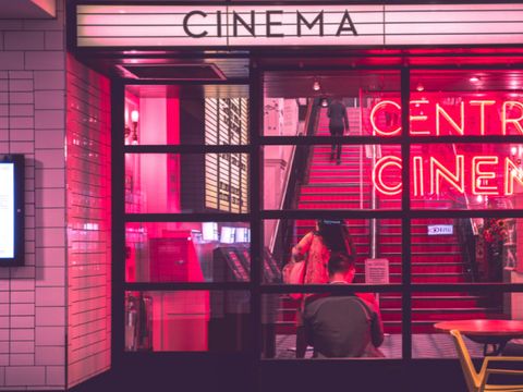 Blick durch Fenster in rot beleuchteten Raum mit vier Personen darin und darüber auf einem Schild das Wort "Cinema"