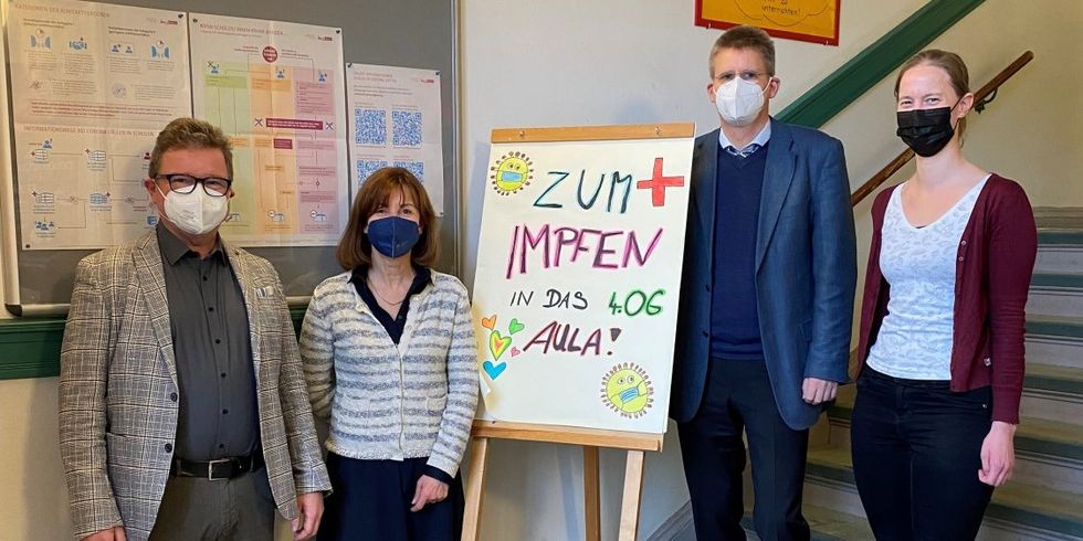 Vier Meschen mit Masken stehen an einer Treppe neben einem Schuld mit der Aufschrift "Zum Impfen in das 4. OG, Aula!"