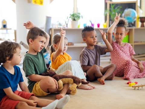 Eine Gruppe Kindergartenkinder sitzt im Zimmer auf dem Boden und hebt die Hände.
