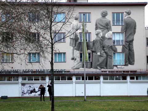 Bildvergrößerung: Auf der Fasse eines großen Blocks sind Menschen in schwarz-weiß abgebildet. Sie wenden sich dem Betrachter mit dem Rücken zu und schauen in die Ferne.