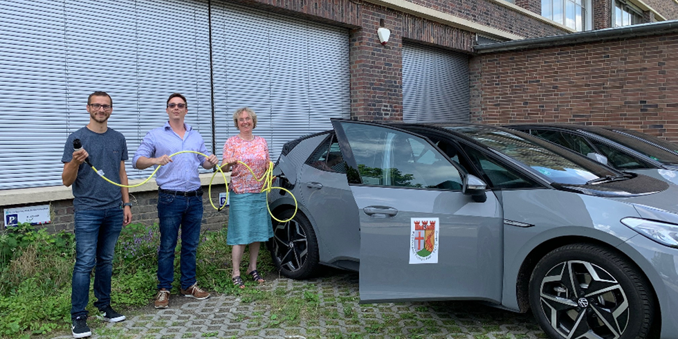 Drei Personen stehen neben drei Neuwagen auf einem Parkplatz und halten das Ladekabel von einem der Elektrowagen.