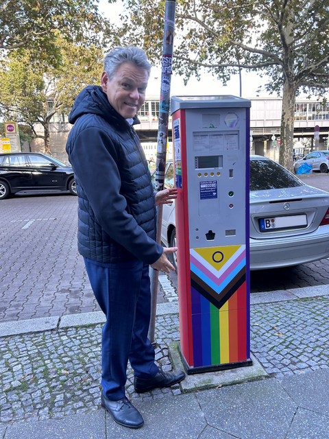 Bildvergrößerung: Ein Mann steht neben einem Parkscheinautomaten, der in der unteren Hälfte mit den Farben der Progress Pride Flagge beklebt ist.