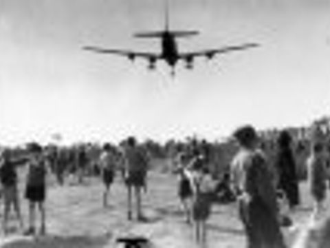 Schwarz-Weiß-Foto eines landenden Flugzeugs über einer Menschenmenge, die zu ihm aufschaut. 