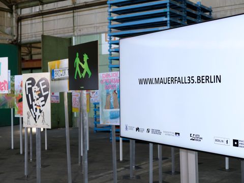 Screen "www.mauerfall35.berlin" mit Freiheits-Schildern im Hintergrund