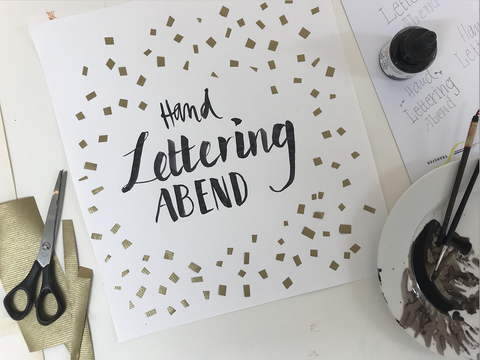 Papierblatt mit handgeschriebenem Text "Hand Lettering Abend" mit goldenen Versierung. Um den Blatt liegen Schere, Pinsel und Farbe.