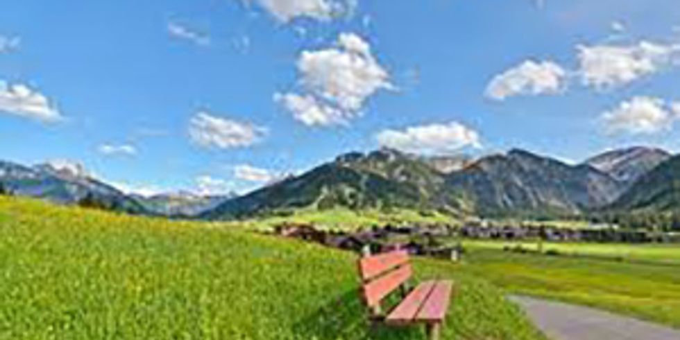 Blick aus dem Tannheimer Tal in Österreich auf die Alpen, im Vordergrund eine Bank auf einer Wiese an einem Weg